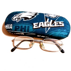 Può includere: Un paio di occhiali in metallo color argento con lenti rettangolari posizionati davanti a una custodia rigida per occhiali color verde acqua e bianco del marchio Philadelphia Eagles, con il logo della squadra e il testo EAGLES e PHI.