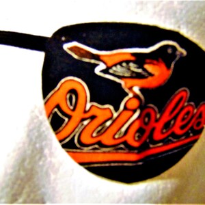 May include: A black eye patch with an orange Baltimore Orioles logo and a bird graphic. The logo says "Orioles" in white lettering.