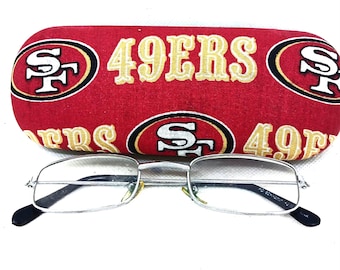 Puede incluir: Estuche para gafas de tela roja y dorada con el logotipo de los San Francisco 49ers y las palabras "49ERS" impresas en él. Un par de gafas con montura plateada están encima del estuche.