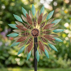 May include: A decorative metal wind spinner with a leaf design. The leaves are a mix of teal, bronze, and gold, arranged in a circular pattern. The wind spinner is mounted on a black pole, set against a blurred garden background.