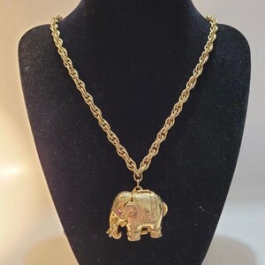 May include: A gold-toned necklace featuring a thick, twisted rope chain and a pendant shaped like an elephant. The elephant pendant has a textured surface with intricate detailing and a small, round pink gemstone set into the side of its head.