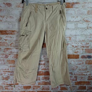 May include: Khaki-colored cargo pants with multiple pockets and zippers. The pants have a relaxed fit and are made of a durable fabric. The pants are hanging on a black hanger against a brick wall.