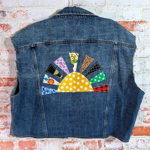 May include: A blue denim vest featuring a large, semi-circular sun applique on the back. The sun has a yellow polka-dot center with radiating triangular rays in various patterns, including floral, leopard print, plaid, and polka-dot fabrics.