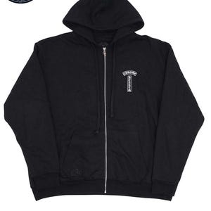 May include: Black zip-up hoodie with a hood and a silver zipper. The hoodie features a small white emblem on the chest and a small logo on the left sleeve. The hoodie is made of a soft material.