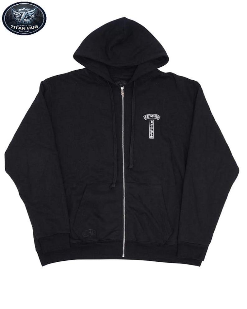 May include: Black zip-up hoodie with a hood and a silver zipper. The hoodie features a small white emblem on the chest and a small logo on the left sleeve. The hoodie is made of a soft material.