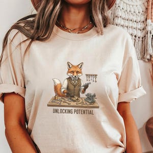 May include: Beige t-shirt featuring a cartoon fox in a vest, holding tools at a workbench. The text "UNLOCKING POTENTIAL" is printed below the fox. The fox is surrounded by keys and tools.