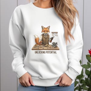May include: White sweatshirt featuring a cartoon fox in a vest, holding a key and working at a table with keys and a small machine. The text "UNLOCKING POTENTIAL" is below the table. The fox has orange and white fur.