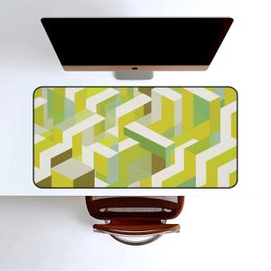 May include: A rectangular desk mat with a geometric pattern in shades of green, white, and brown. The mat is positioned under a computer monitor on a white desk, with a brown office chair visible below.