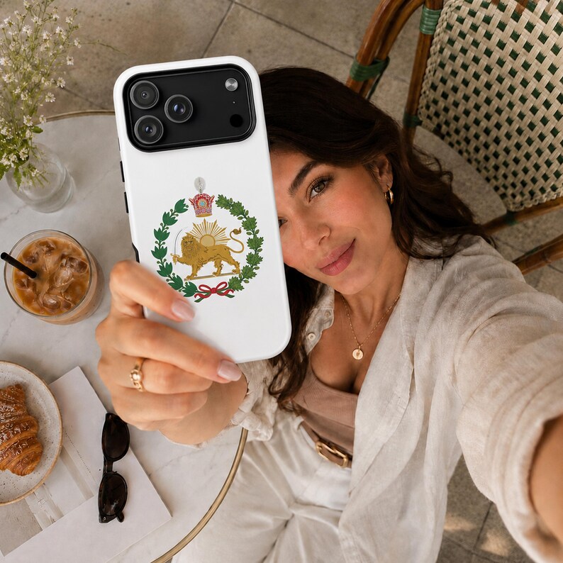 Puede incluir: Una funda de smartphone blanca con un gr&aacute;fico central del emblema del Le&oacute;n y el Sol, que representa un le&oacute;n dorado sosteniendo una espada dentro de una corona de laurel verde, rematado con una corona, sobre un fondo blanco liso.