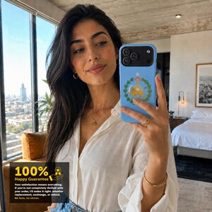 Puede incluir: Una funda de smartphone de color azul claro con un emblema central de un le&oacute;n dorado rodeado por una corona de laurel verde y un icono de corona roja en la parte superior. La funda tiene un recorte negro para el m&oacute;dulo de la c&aacute;mara.