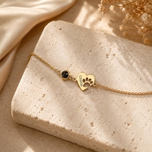 Op de afbeelding: Een goudkleurige kettingarmband met een kleine ronde zwarte steenbedel en een hartvormige hanger gegraveerd met de naam Charlie en een uitgesneden pootafdruk. De fijne metalen ketting verbindt beide bedels tot een eenvoudig sieraad.