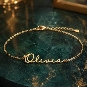 Op de afbeelding: Een goudkleurige armband met de naam "Olivia" in een sierlijk lettertype. De armband ligt op een donkergroen marmeren oppervlak. De ketting is fijn en heeft een kleine sluiting.
