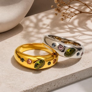 Op de afbeelding: Twee ringen, één goudkleurig en één zilverkleurig, worden getoond. De gouden ring heeft een grote ovale groene steen en kleinere roze en groene stenen. De zilveren ring heeft een vergelijkbaar ontwerp met blauwe en roze stenen. Beide ringen liggen op een neutrale ondergrond.