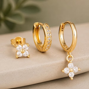 Puede incluir: Tres pendientes dorados. Uno es un pendiente de botón con un diseño de flor de cuatro pétalos, otro es un aro pequeño con piedras transparentes, y el tercero es un aro con un diseño de flor de cuatro pétalos colgante con piedras transparentes.