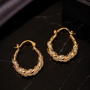 Puede incluir: Un par de pendientes de aro dorados con un diseño de cuerda trenzada. Los aros están incrustados con pequeños cristales brillantes. Los pendientes se exhiben sobre una superficie de mármol negro.