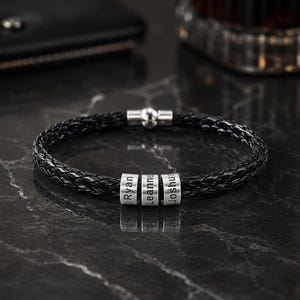 Könnte beinhalten: Ein schwarzes geflochtenes Lederarmband mit drei zylindrischen silberfarbenen Metallperlen. Jede Perle ist mit einem Namen graviert: Ryan, Leanne und Joshua. Das Armband wird mit einem polierten silberfarbenen Magnetverschluss geschlossen.