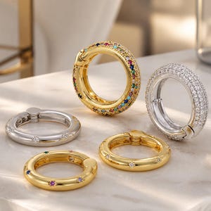 Könnte beinhalten: Fünf Ringe in Gold- und Silbertönen. Zwei goldene Ringe sind mit bunten Edelsteinen und sternförmigen Akzenten verziert. Ein silberner Ring ist mit kleinen, klaren Steinen besetzt. Die anderen beiden Ringe sind silber- und goldfarben mit klaren Steinakzenten.