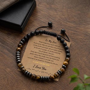 Peut inclure: Un bracelet de perles alternant des perles noires mates, des perles œil-de-tigre et des entretoises métalliques couleur hématite. Le bracelet repose sur une carte rectangulaire avec le texte To My Son, un message inspirant et du code Morse.