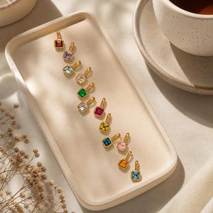 Puede incluir: Una colección de colgantes dorados con piedras cuadradas de varios colores, incluyendo rojo, morado, verde y azul. Los colgantes están dispuestos sobre una bandeja de color claro, con una taza de té en el fondo.