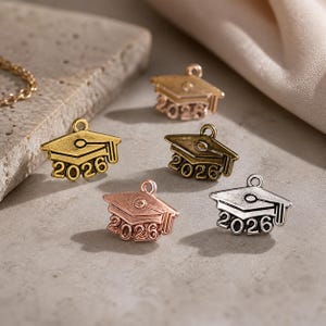 Può includere: Cinque ciondoli a forma di cappello di laurea nei toni oro, bronzo, rame e argento. Ogni ciondolo presenta l'anno "2026" e un anello per attaccarlo a una collana o un braccialetto. I ciondoli sono sparsi su una superficie neutra.