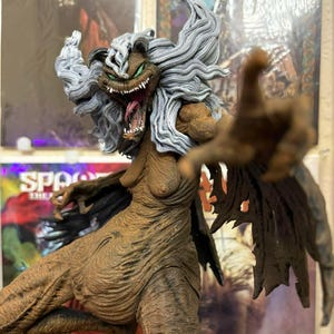 May include: A detailed figurine of a monstrous female character with gray hair, green eyes, and an open mouth revealing sharp teeth. The figure has a textured, brown body and outstretched arms, with dark wings. Comic book covers are in the background.