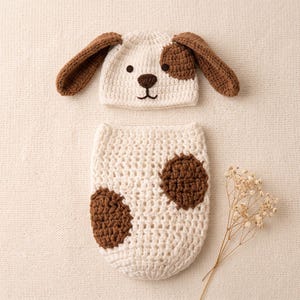 Pode incluir: Um conjunto de crochê composto por um gorro cor de creme com orelhas longas castanhas e um design de cara de cão, combinado com um saco de casulo de crochê a condizer em cor de creme com duas grandes manchas circulares castanhas à frente.