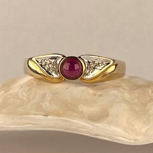 Op de afbeelding: Een gouden ring met een centrale ronde cabochon geslepen donkerroze edelsteen in een zetting. De ringband bevat aan weerszijden van de middelste steen twee driehoekige zilverkleurige secties, elk versierd met drie kleine ingebedde heldere steentjes.