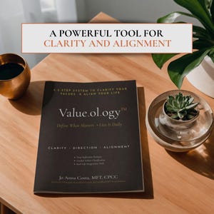 May include: A book titled "Value.ology" with the subtitle "Define What Matters • Live It Daily" on a wooden table. The book's cover is dark with white text. The text "A POWERFUL TOOL FOR CLARITY AND ALIGNMENT" is at the top. A succulent plant in a glass bowl is to the right.
