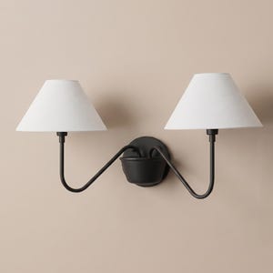 May include: A double wall sconce featuring a matte black metal base and two curved arms extending outward. Each arm supports a white, conical fabric lampshade. The fixture is designed for wall mounting with a central circular backplate.