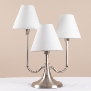 May include: A three-light table lamp featuring a brushed nickel finish base with three curved arms of varying heights. Each arm supports a white, tapered fabric lampshade. The central lamp is positioned lower than the two outer lamps.