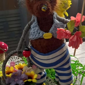 May include: A brown fuzzy creature figurine with white hair, wearing a light blue vest and blue and white striped pants. It holds a small basket filled with yellow and purple flowers, a ladybug charm, and a stem with pink flowers.