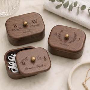 May include: Three rectangular dark wood boxes with rounded corners and brass knobs. One box is open, revealing white contents. The lids feature laser-engraved designs including initials K and W, A and J, and mountain motifs with the text Adventure Together.