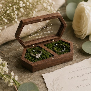 May include: A hexagonal dark wood ring box with a clear acrylic lid featuring a white mountain, moon, and star graphic with the text Charlotte & William 05.25.2025. The interior contains two compartments filled with green moss holding two silver rings.
