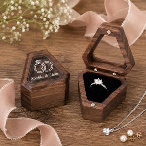 May include: Two hexagonal dark wood ring boxes with hinged lids. One box is closed, featuring a white graphic of two interlocking rings and the text "Sophia & Liam". The open box contains a silver ring with a clear stone. A silver necklace and earrings are nearby.