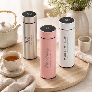 May include: Three stainless steel insulated water bottles with digital temperature displays. One is silver with an "M" and text, another is pink with "Kevin Chang" and text, and the third is white with "Jackson Williams" and text.