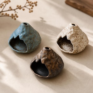 May include: Three ceramic decorative pieces in the shape of small mountains. One is blue, one is brown, and one is off-white. Each has a hole at the top and a large opening on one side. The pieces have a textured, rustic appearance.