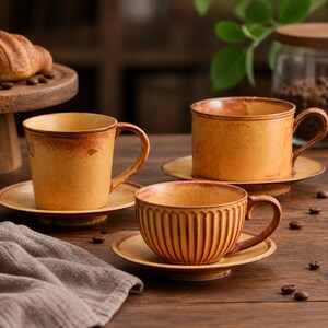 Puede incluir: Tres tazas de café y platillos de cerámica en tonos marrones y beige. Una taza tiene estrías verticales, mientras que las otras son lisas. Las tazas están sobre una mesa de madera con granos de café esparcidos.