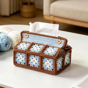 May include: A rectangular tissue box cover crafted from crocheted yarn in brown, light blue, and white. The design features a series of square floral motifs and a roof-like top secured with two small, round, light brown wooden buttons.