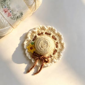May include: A small, cream-colored crocheted hat ornament featuring a scalloped brim, a brown braided ribbon tied in a bow, and a single yellow sunflower accent. The hat is crafted with intricate yarn stitches and has a rounded crown.