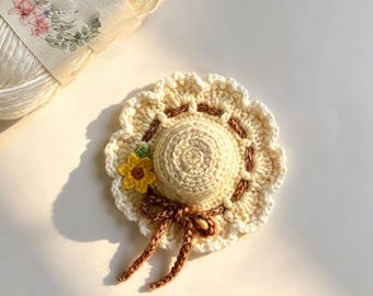 May include: A small, cream-colored crocheted hat ornament featuring a scalloped brim, a brown braided ribbon tied in a bow, and a single yellow sunflower accent. The hat is crafted with intricate yarn stitches and has a rounded crown.