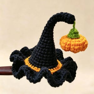 May include: A crocheted black pointed hat with a ruffled brim and an orange band at the base. A small orange pumpkin with a green stem hangs from the tip of the hat by a short green thread. The item is attached to a dark brown wooden stick.