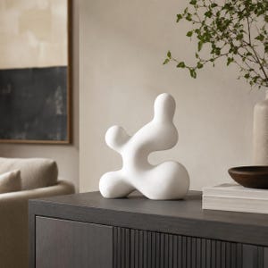 Peut inclure: Une sculpture abstraite blanche avec une finition mate lisse et des formes organiques, arrondies et fluides. La pièce présente plusieurs formes bulbeuses interconnectées qui créent une silhouette continue et fluide, formant un objet décoratif unique.