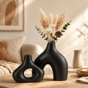 Pode incluir: Dois vasos de cerâmica preto fosco com formas orgânicas e assimétricas. O vaso mais alto apresenta um arco central e contém um arranjo de flores secas, incluindo erva-das-pampas, folhas de eucalipto e uma pequena pinha.