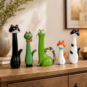 May include: A set of five cat figurines with elongated necks and distinct features. The collection includes a solid black cat, a green cat with a red scarf, a green cat with a curled tail, a small white cat with orange ears, and a white cat with black spots.