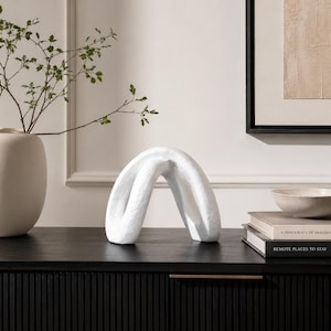 May include: A white sculptural object with a curved, arching form and a textured surface. The piece features a continuous, folded shape that creates an open, hollow centre, resting on a flat base. The sculpture is matte white in colour.