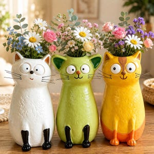 May include: Three cat-shaped ceramic vases in white, light green, and yellow, each featuring wide eyes, whiskers, and black or orange painted paws. The vases are filled with arrangements of white daisies, pink roses, and small blue and purple flowers.