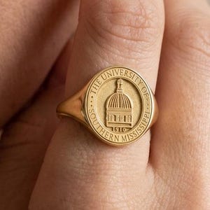 May include: A gold signet ring featuring the seal of The University of Southern Mississippi. The ring has a round face with the university's emblem and the year 1910. The ring is worn on a finger.