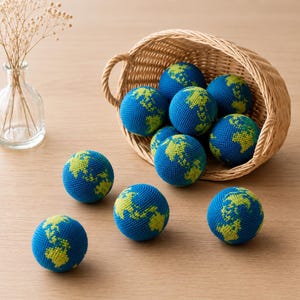 May include: A collection of blue and green crocheted globe-shaped balls, some spilling from a woven basket. The balls feature a detailed representation of the Earth's continents in green against a blue background. The scene is set on a light-colored wooden surface.