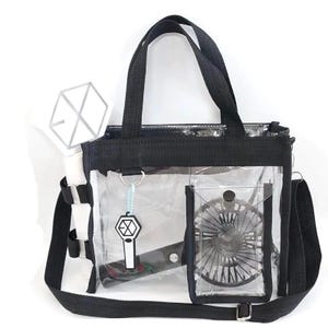 Puede incluir: Bolso tote transparente con ribete, asas y bandolera negros. El bolso tiene un pequeño bolsillo que contiene un ventilador negro. En el interior hay un palo de luz blanco y un llavero con un logotipo.