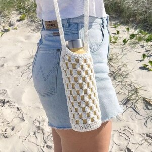 May include: A white crochet water bottle holder with a shoulder strap. The holder has a checkered pattern and contains a yellow water bottle. The background is a sandy beach with green plants.